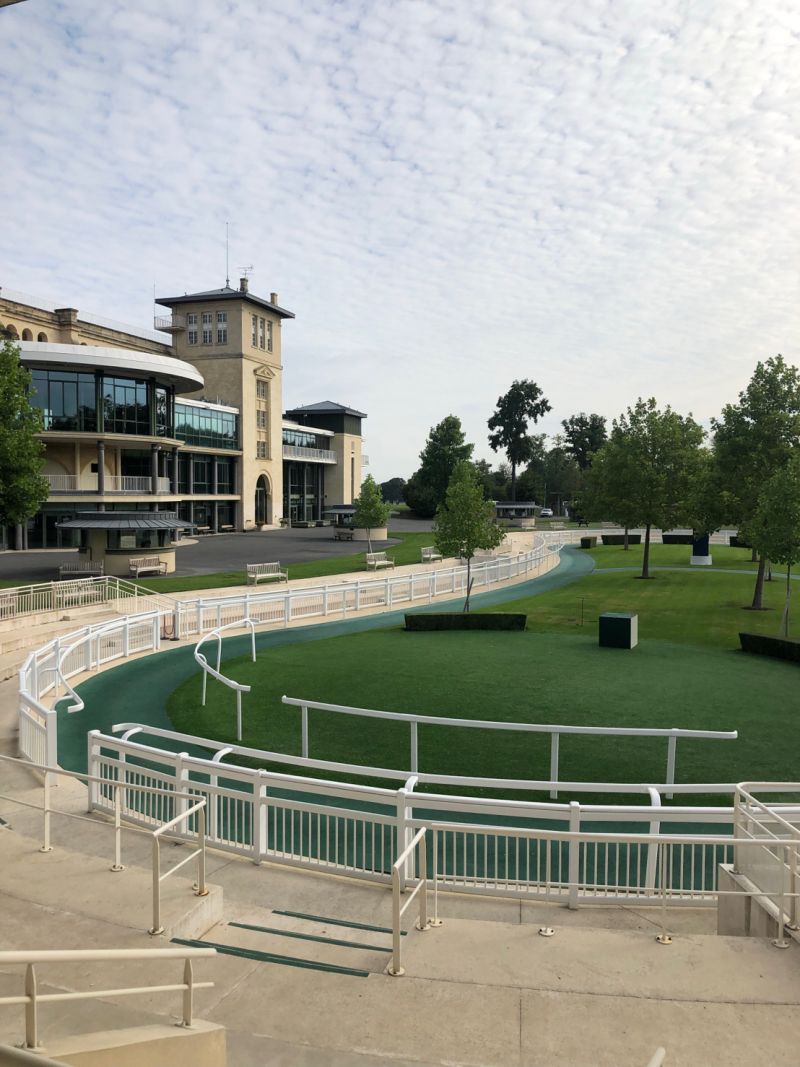 Empty horse racing paddock with white railings and a tall building with large windows in the background under a cloudy sky.