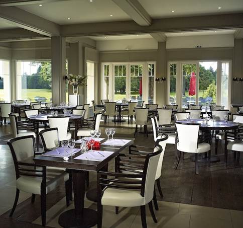 Elegant empty restaurant dining area with white cushioned chairs, dark wood tables set with napkins and glassware, and large windows overlooking a green outdoor space.