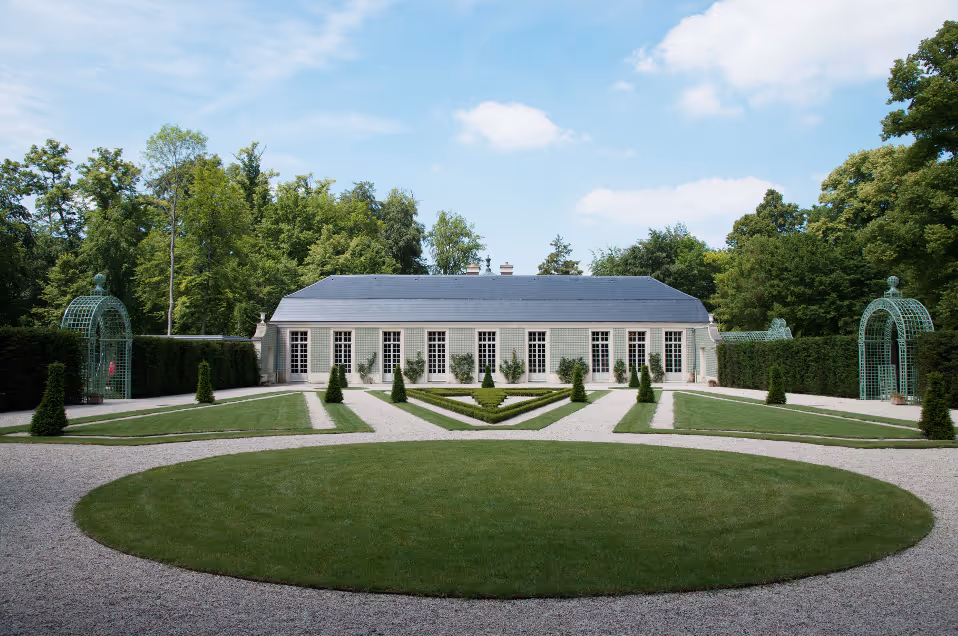 Symmetrical garden with manicured grass, gravel pathways, and arched green trellises leading to a long building with large windows.