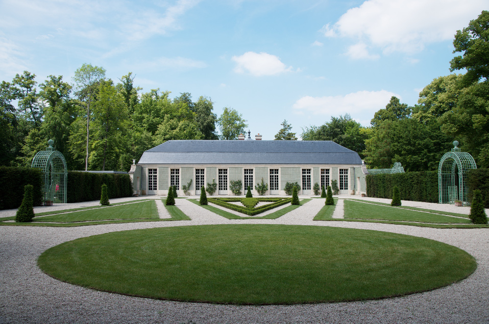 Symmetrical garden with manicured grass, gravel pathways, and arched green trellises leading to a long building with large windows.