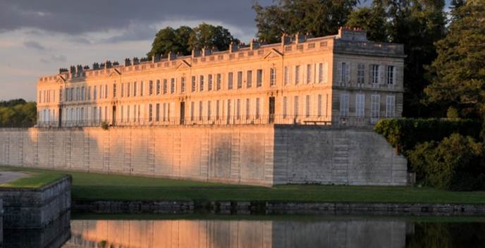 Historic stone building illuminated by warm sunlight, reflected in a calm body of water with surrounding greenery.