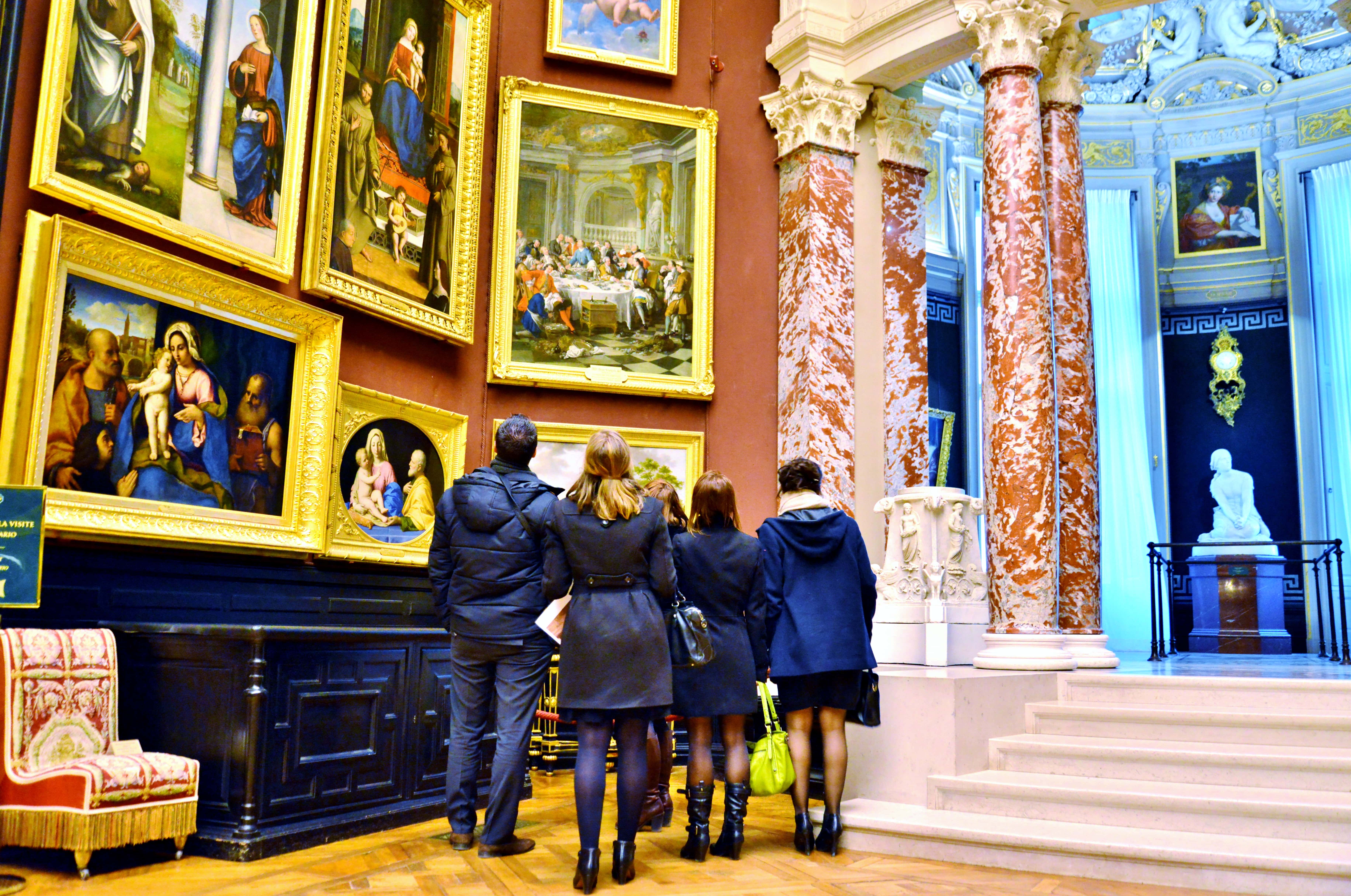 Group of five people viewing classical paintings in a museum with ornate red and white marble columns and a white statue in the background.
