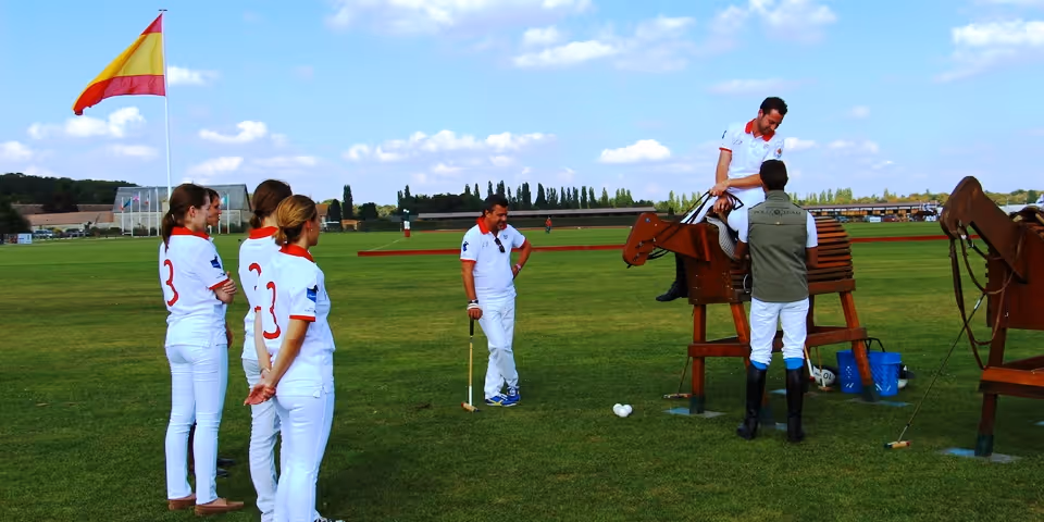 Group of polo players in white uniforms standing on grass near wooden horse structures, with one player sitting on a wooden horse.
