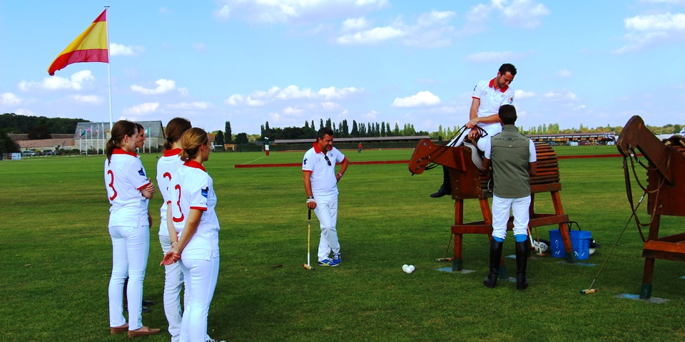 A group of people in white polo attire standing on a grassy field with wooden practice horses and polo mallets.