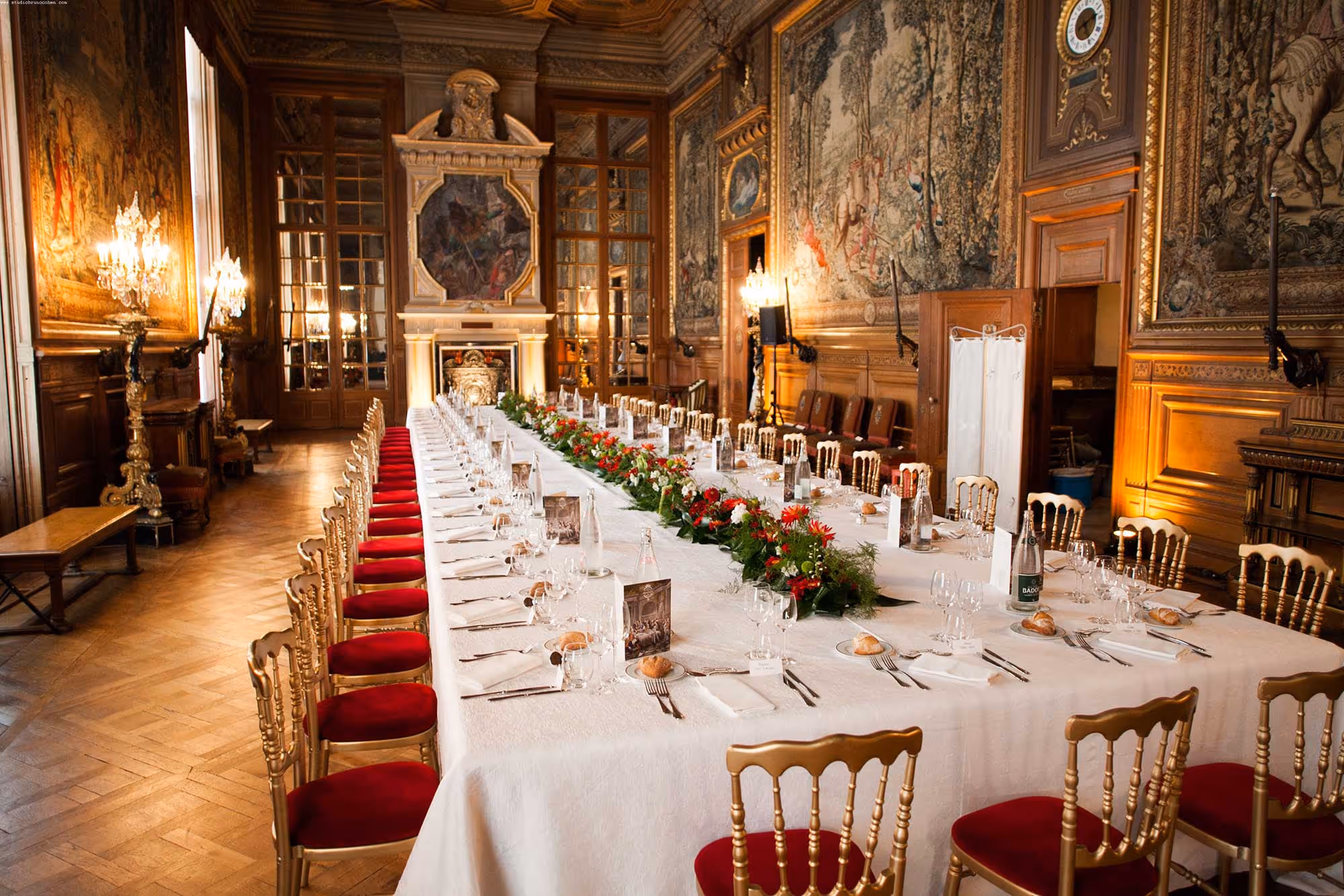 Elegant long dining table set with white tablecloth, red floral centerpieces, and gold chairs with red cushions in a richly decorated historic room with tapestries and chandeliers.