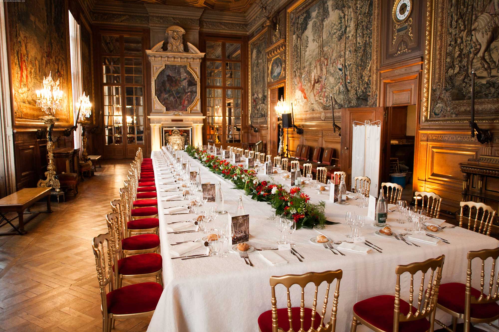 Elegant long dining table set with white tablecloth, red floral centerpieces, and gold chairs with red cushions in a richly decorated historic room with tapestries and chandeliers.