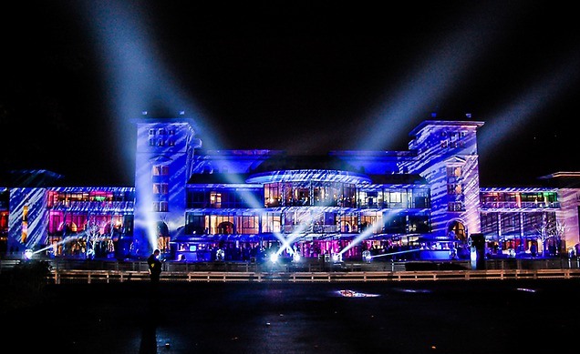 Large building illuminated with colorful blue and purple lights and beams at night.
