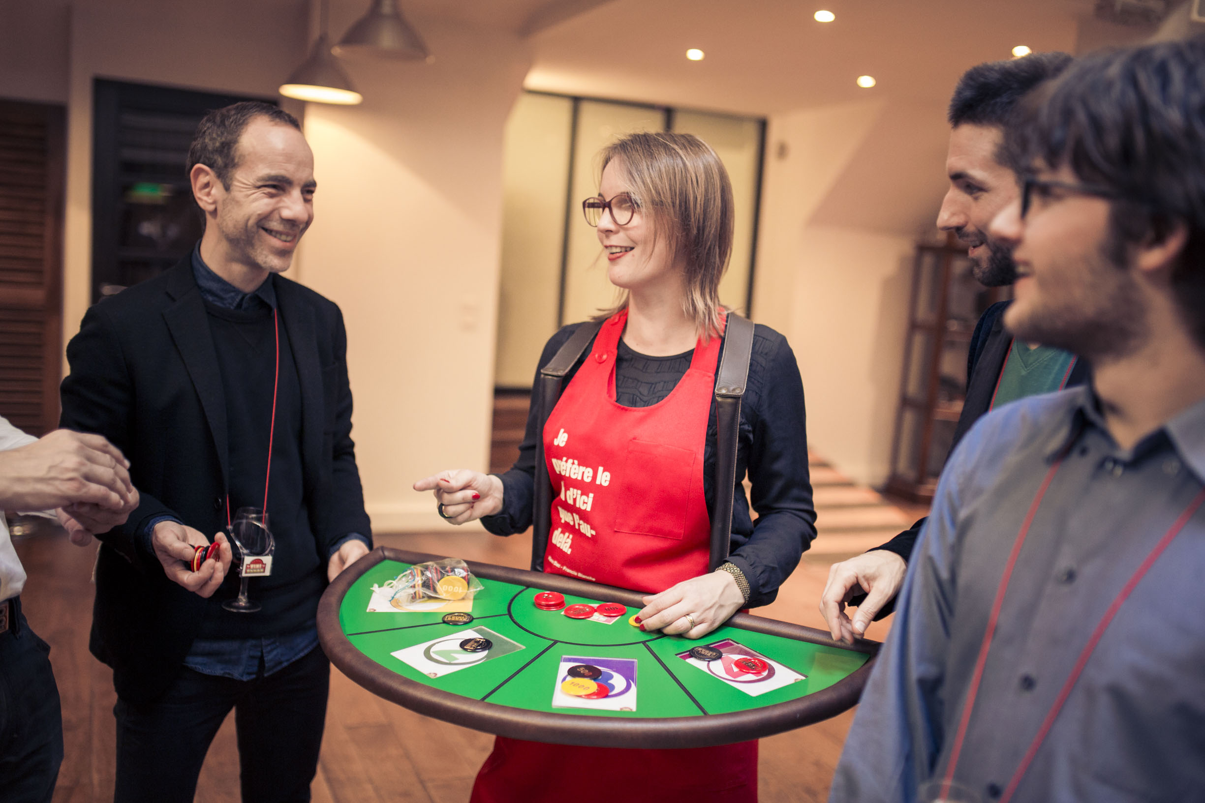 Woman wearing glasses and a red apron holding a green gaming tray with chips, surrounded by smiling men at an indoor event.