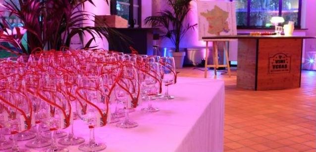 Table covered with a white cloth holding numerous empty wine glasses with red ribbons, set in a room with purple lighting and plants.