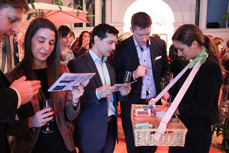 People dressed in business attire examining brochures and holding drinks at an indoor event, while a woman with a basket on a strap offers items.