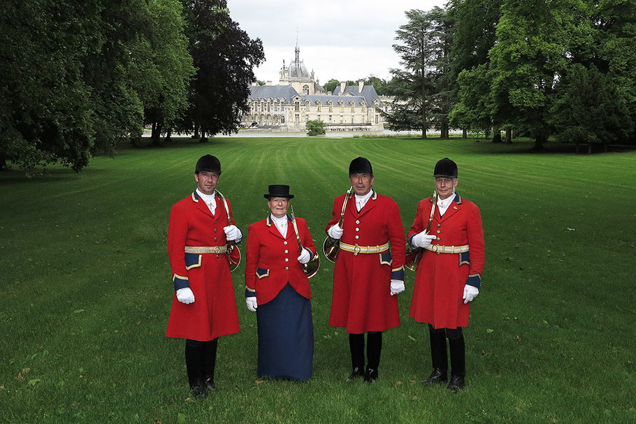 Four people in traditional red hunting coats standing on a green lawn with a large chateau in the background.