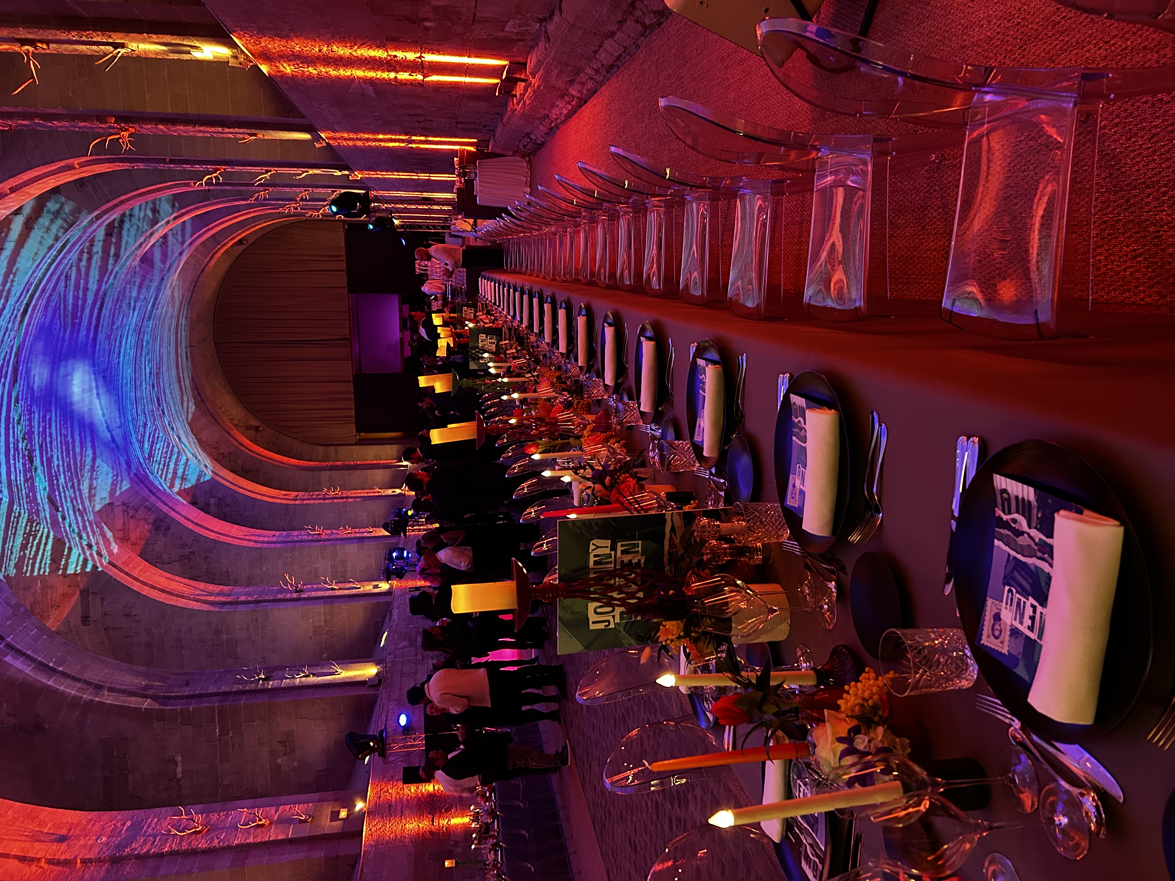 Long banquet table set with black plates, white napkins, candles, and floral arrangements in a large arched hall with purple and orange lighting and clear chairs.