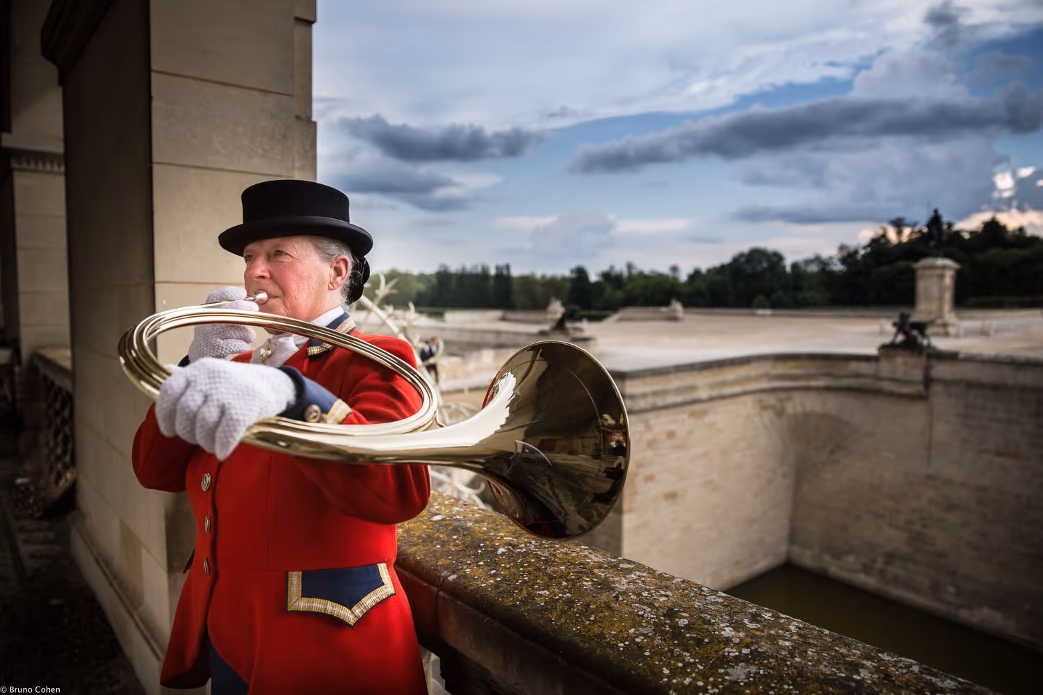 Person in red ceremonial uniform and black hat playing a brass horn outdoors near a stone railing and historic building.