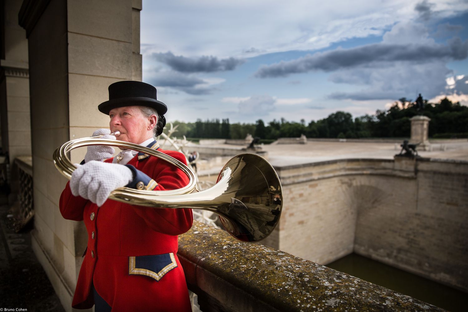 Person in red ceremonial uniform and black hat playing a brass horn outdoors near a stone railing and historic building.