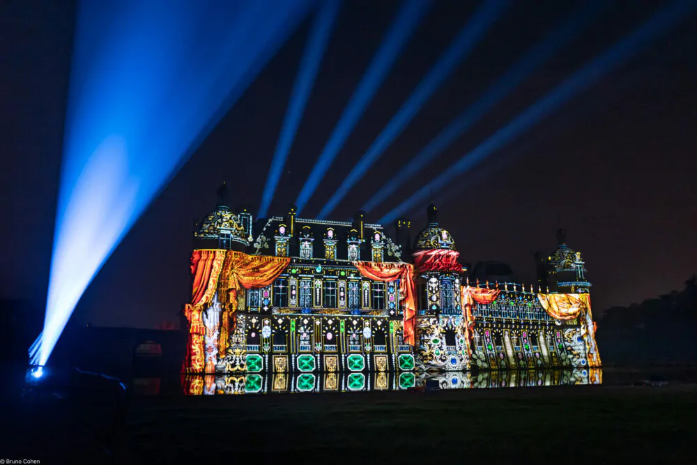 Night view of a historic château illuminated with colorful light projections and blue spotlights shining into the sky.