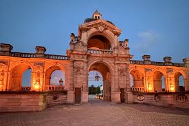 Historic stone archway and surrounding structure illuminated with warm lights under a dusk sky.