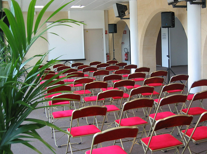 Conference room with rows of red cushioned folding chairs facing a blank projection screen and speakers.