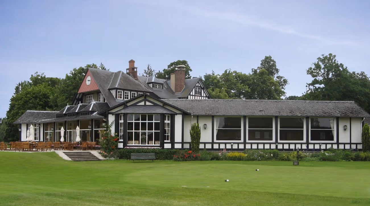 Large traditional clubhouse building with outdoor seating on a green golf course under a clear sky.