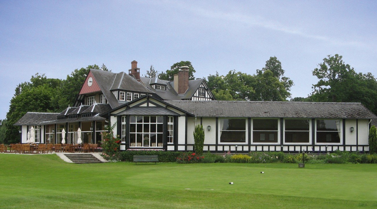 Traditional-style clubhouse building with large windows and outdoor seating overlooking a green golf lawn.