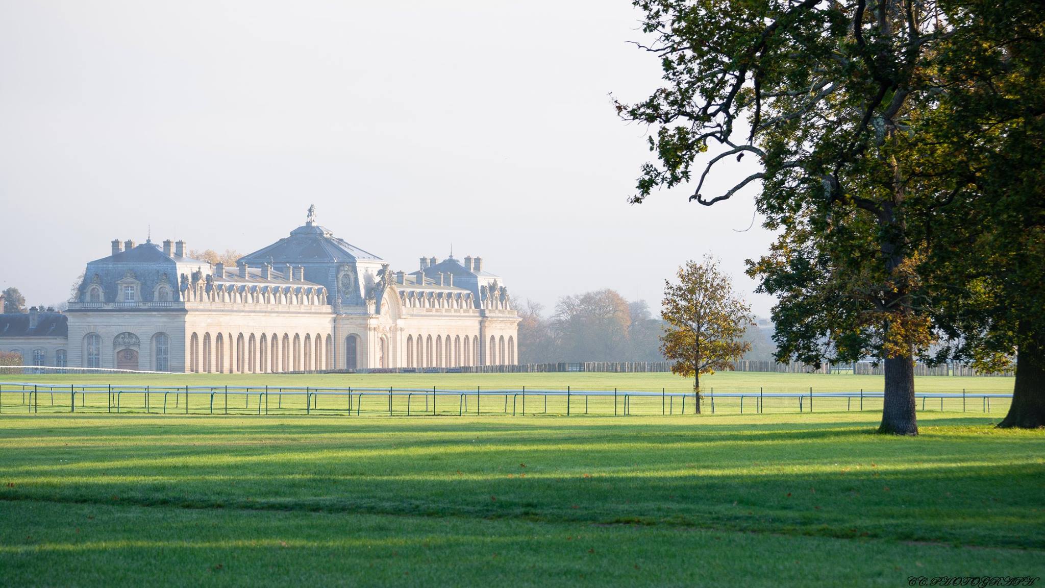 Large historic stone building with arched windows viewed across a green lawn with trees casting shadows.