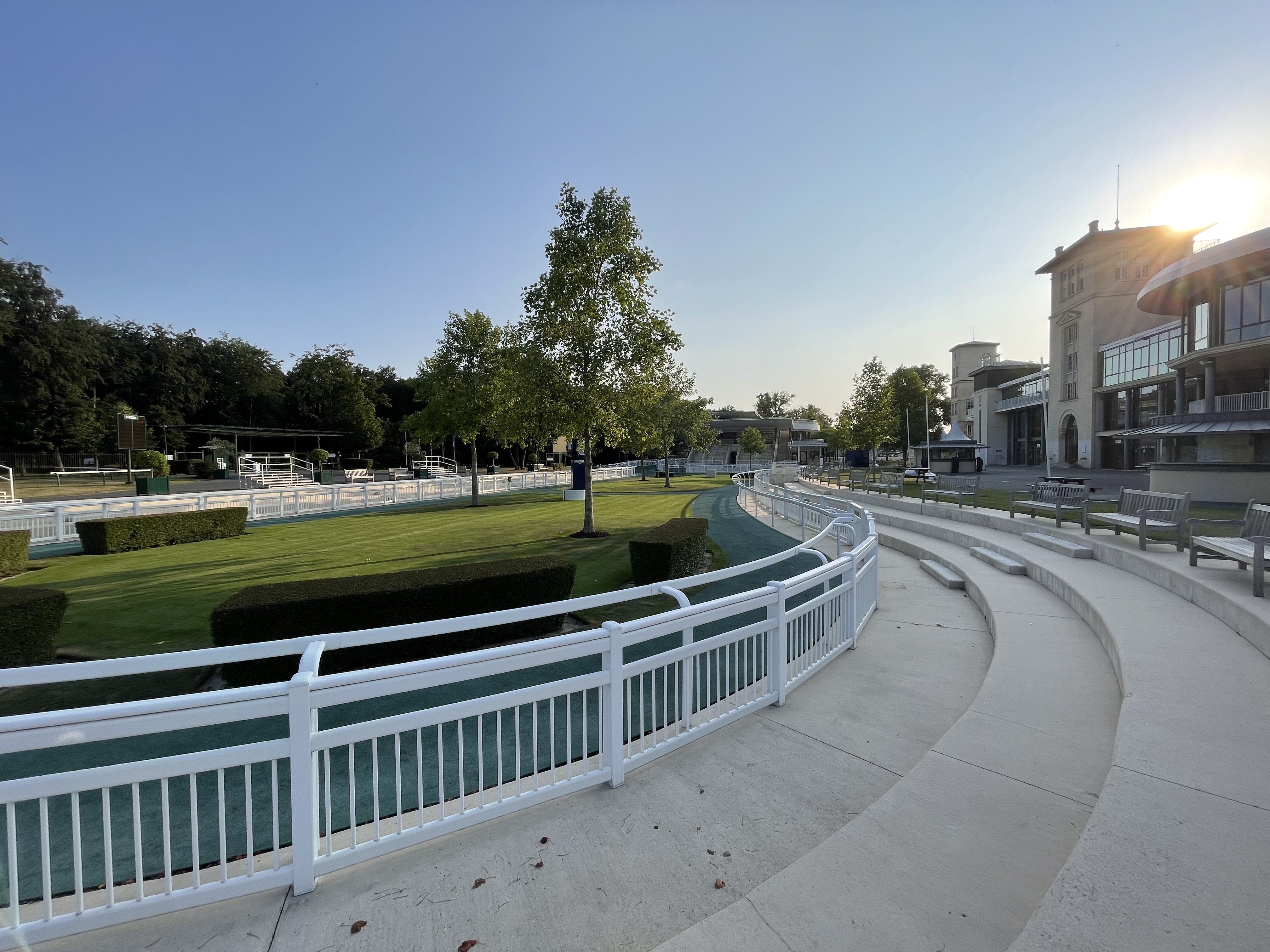Empty racetrack with curved white fencing, green grass, trees, and tiered seating under a clear sky with the sun setting behind buildings.