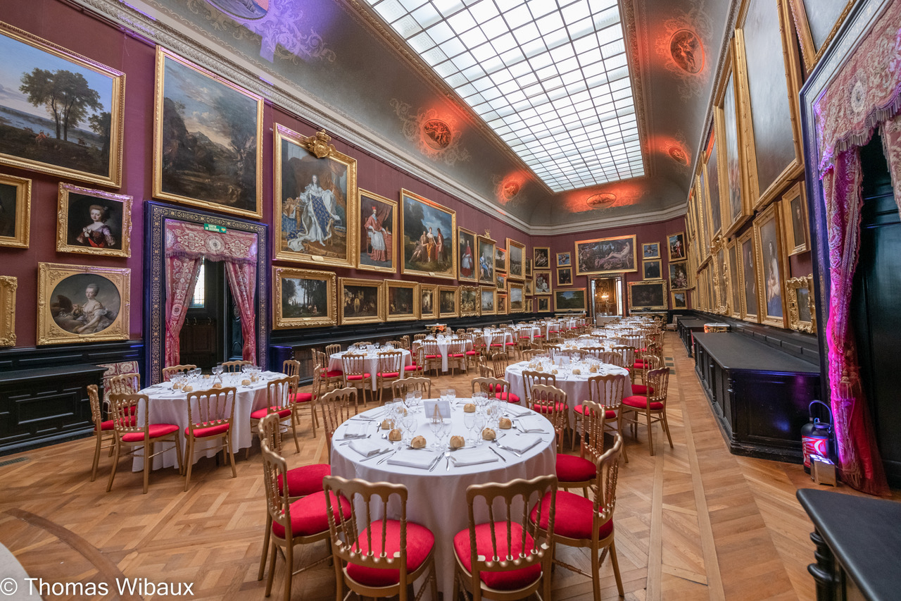 Elegant banquet room with round tables set for dining surrounded by red cushioned gold chairs, ornate paintings covering purple walls, and a large skylight ceiling.