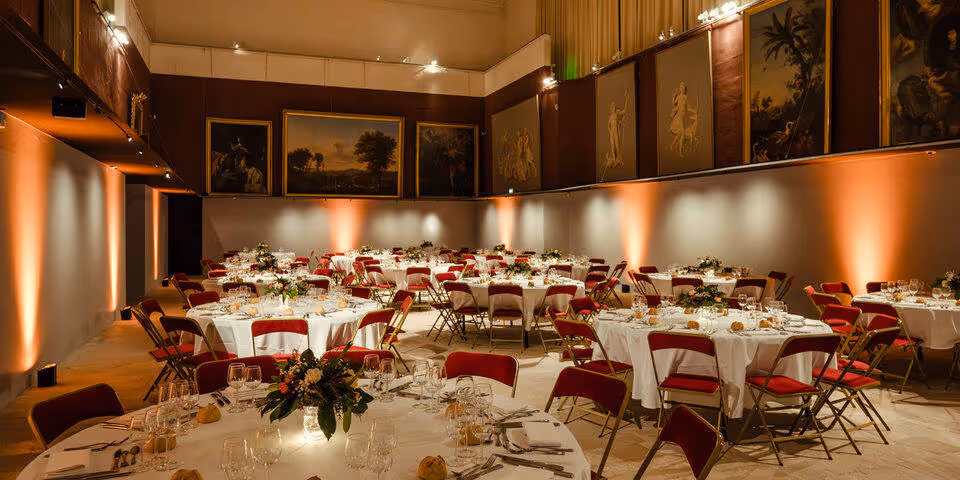 Elegant banquet hall with round tables set with white tablecloths, red chairs, glassware, and floral centerpieces under warm lighting and framed paintings.