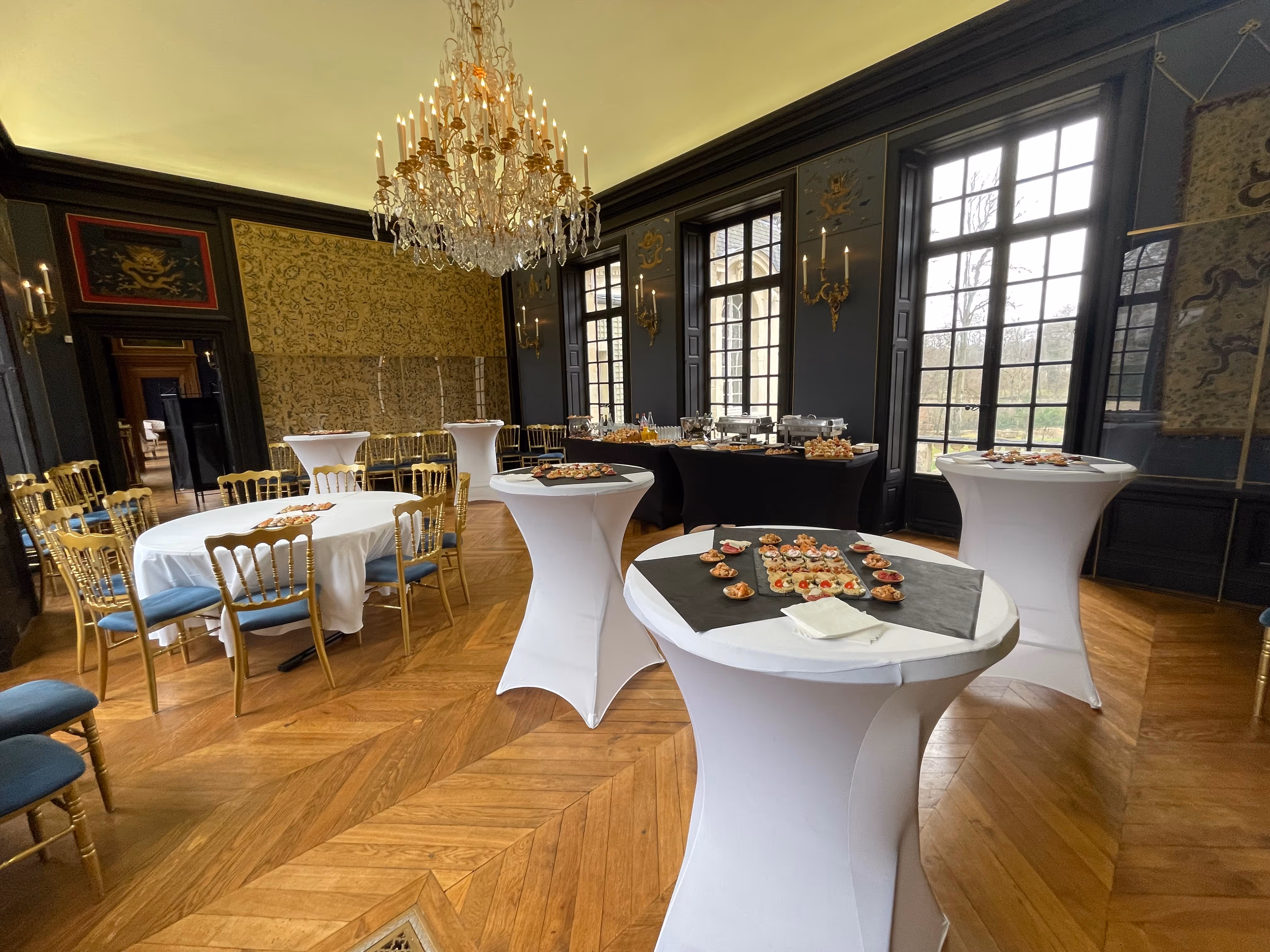 Elegant banquet room with wooden floors, gold chairs, large windows, white tablecloth-covered round tables, and a crystal chandelier.