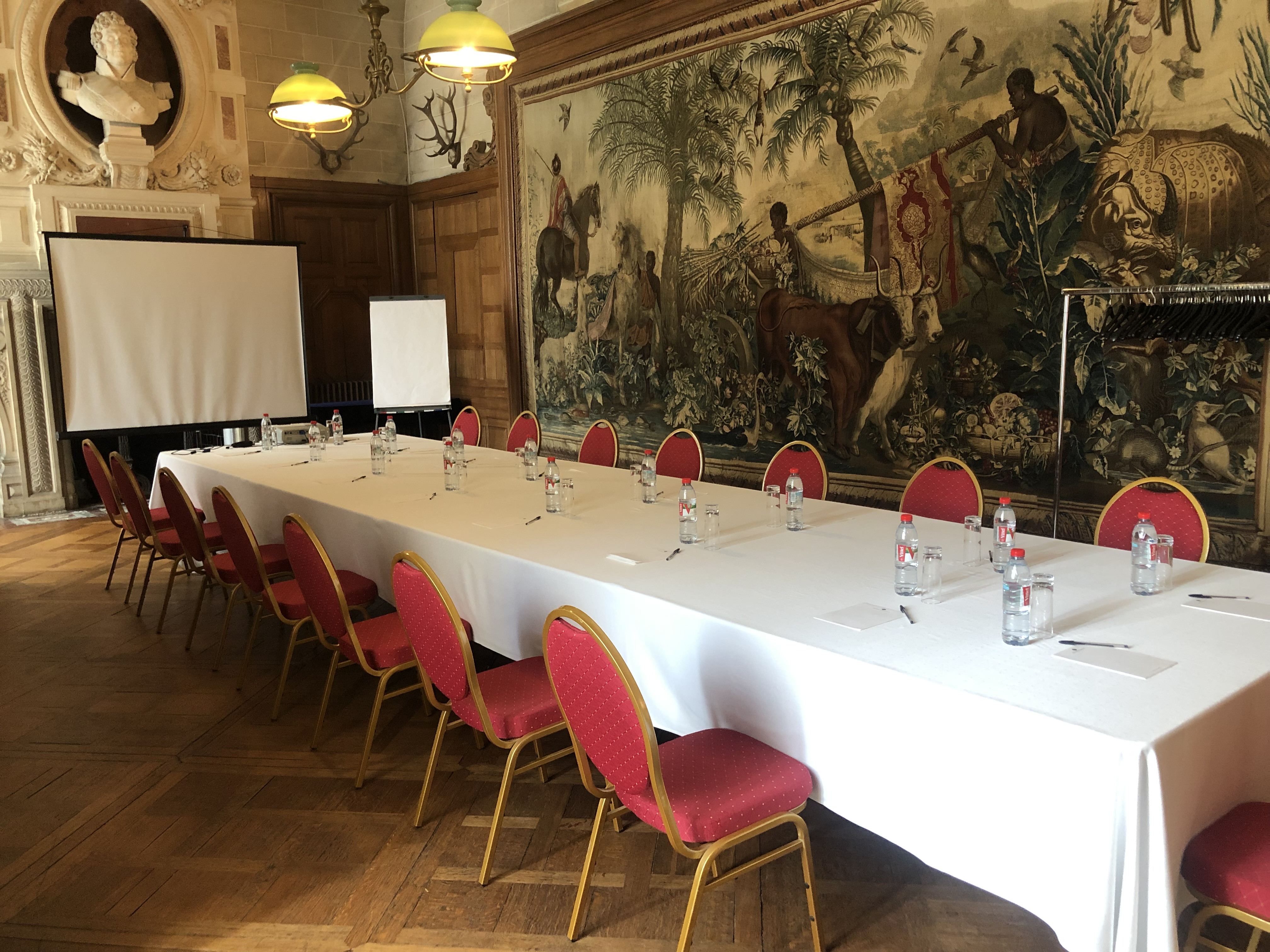 Long conference table with red chairs, water bottles, notepads, and pens in an ornate room with a large tapestry and classical bust.