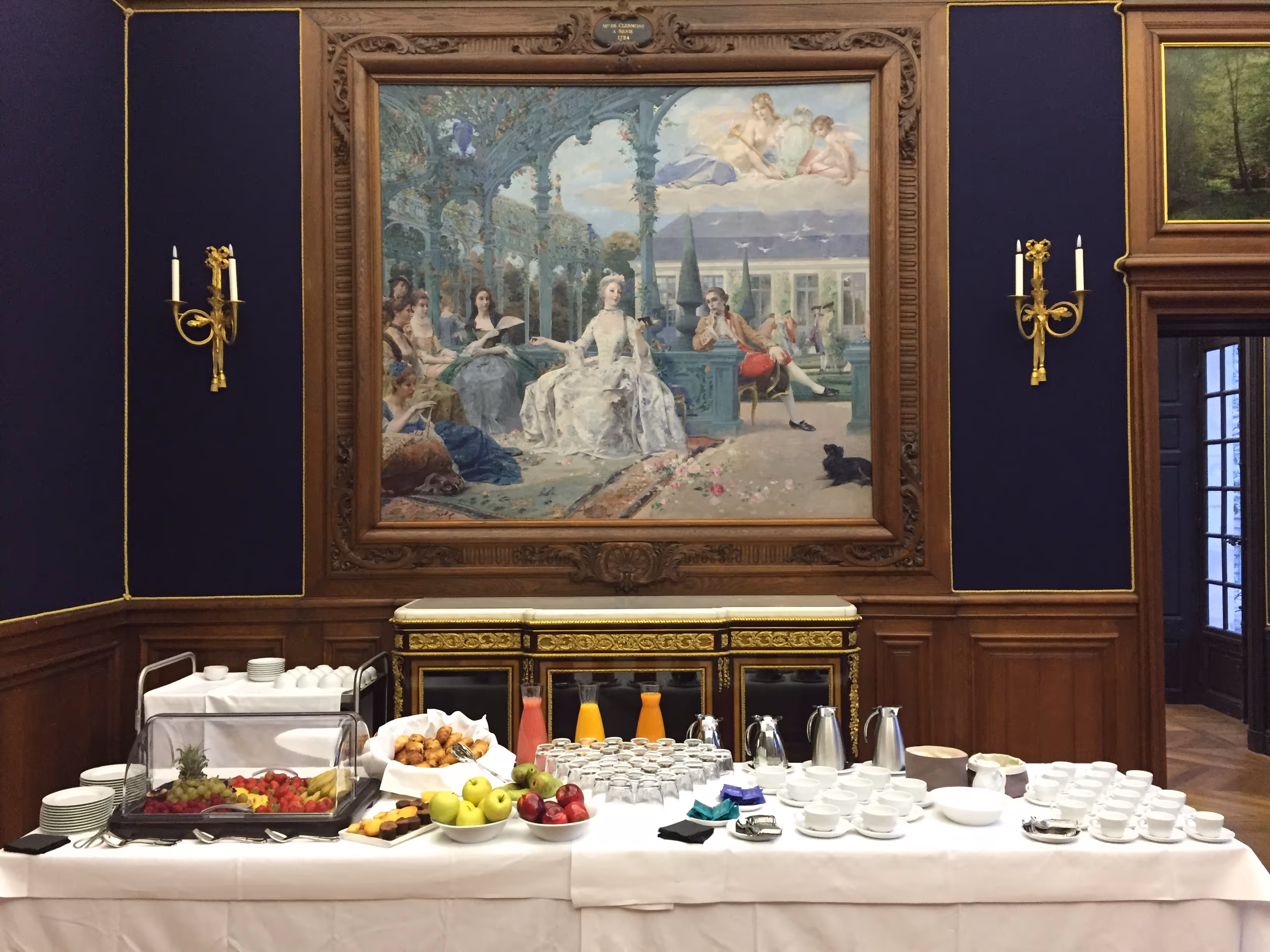 Buffet table with fruit, pastries, juice, cups, and coffee pots set in a room with wooden paneling and a large classical painting on the wall.
