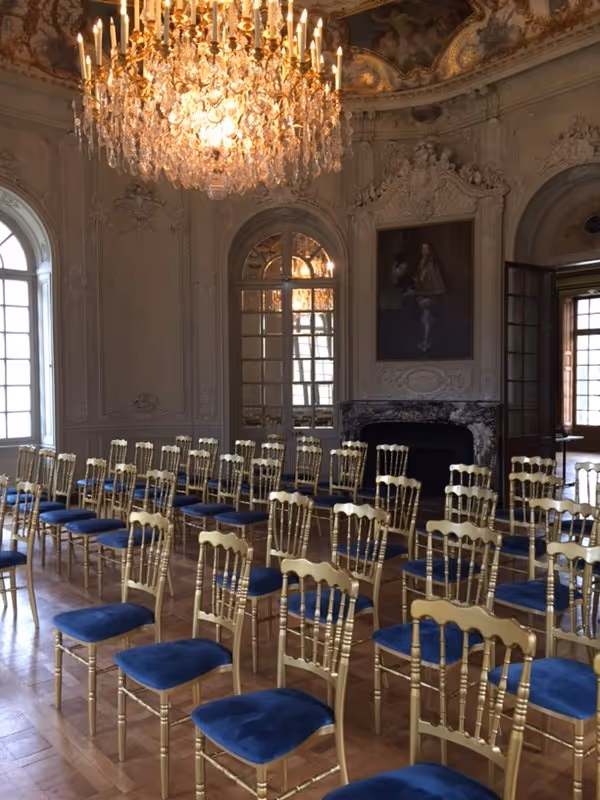 Elegant room with rows of gold chairs with blue cushions, a large crystal chandelier, ornate ceiling, and a fireplace with a portrait above it.