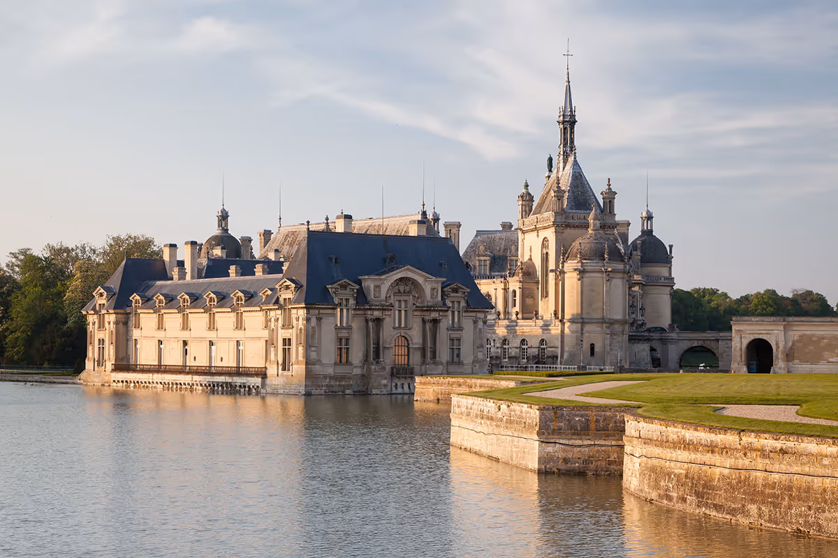 Historic château with ornate towers surrounded by a water moat and green lawns under a partly cloudy sky.