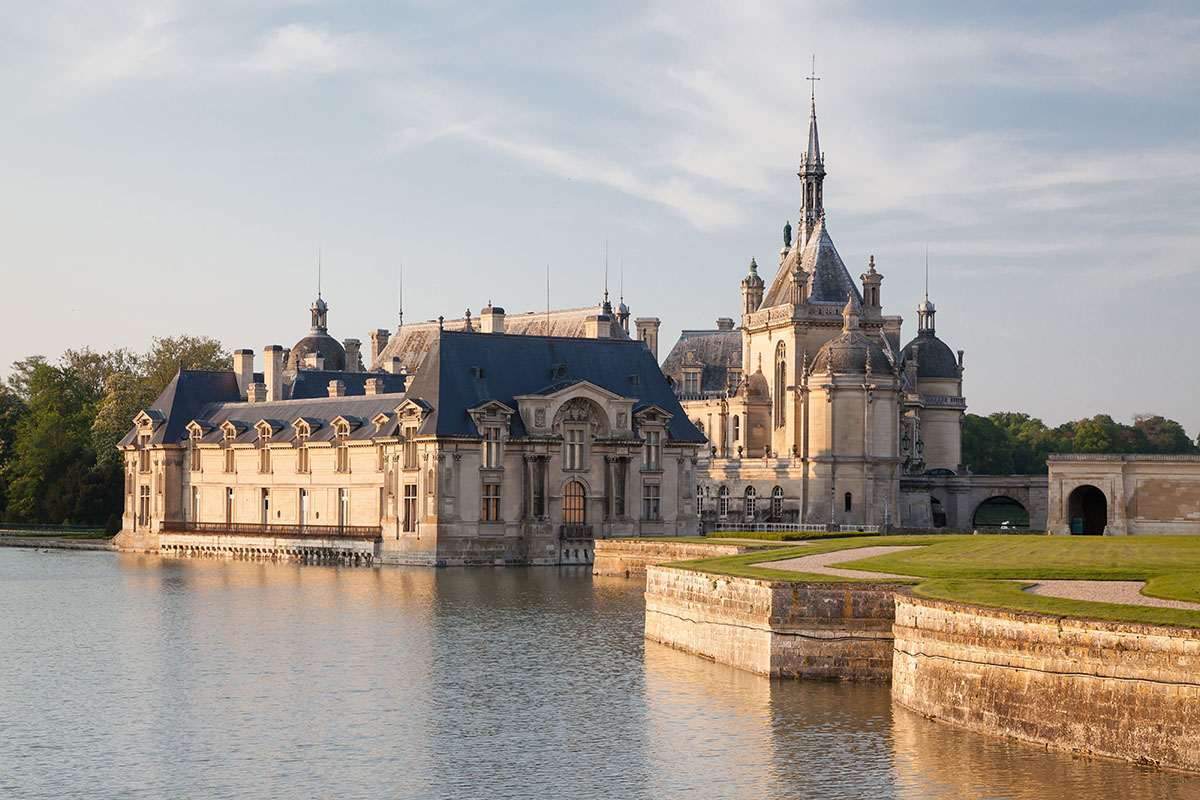 Historic château with ornate towers surrounded by a water moat and green lawns under a partly cloudy sky.