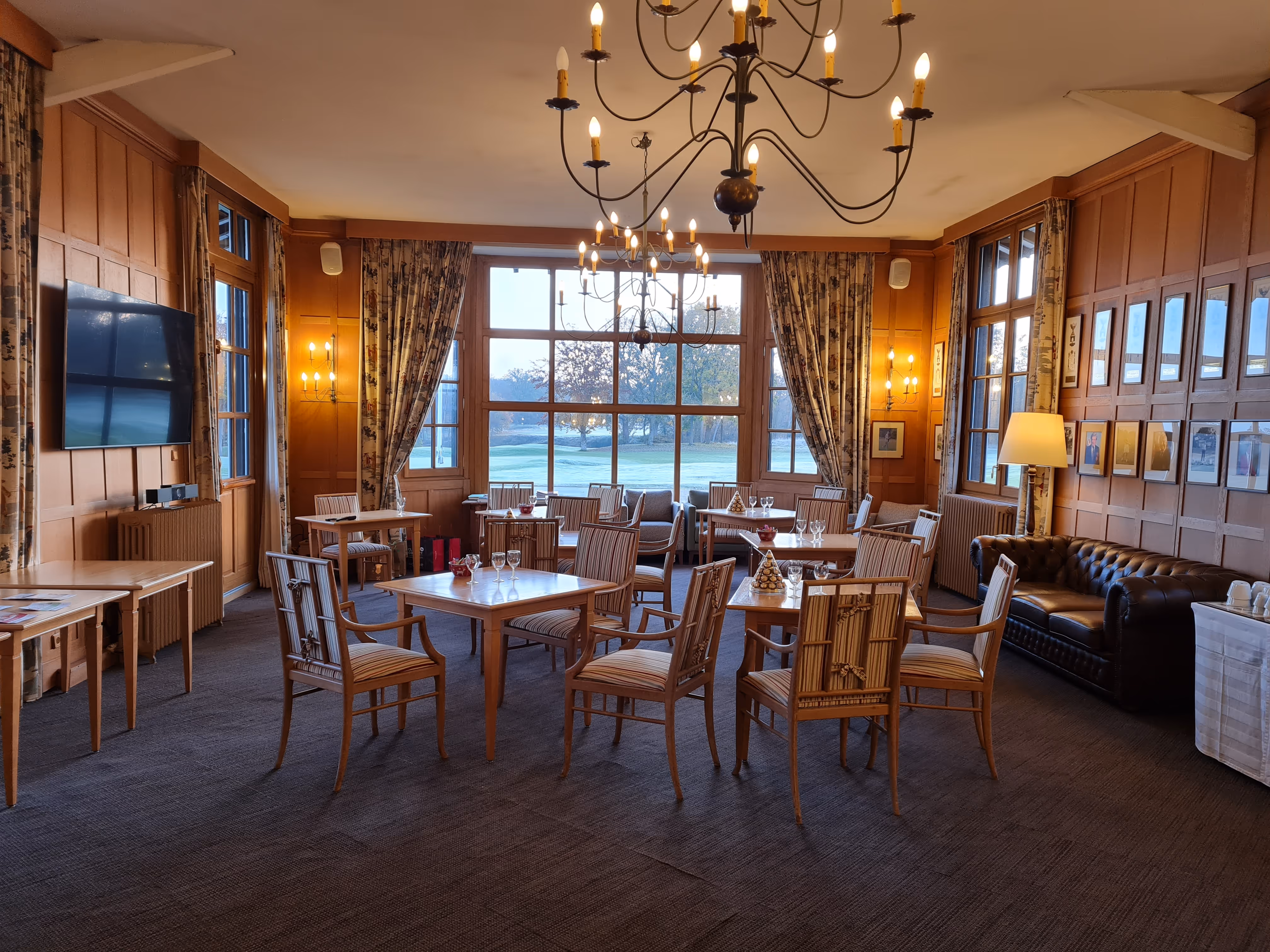 Elegant dining room with wooden panel walls, chandeliers, and tables with chairs arranged near large windows overlooking a green outdoor area.