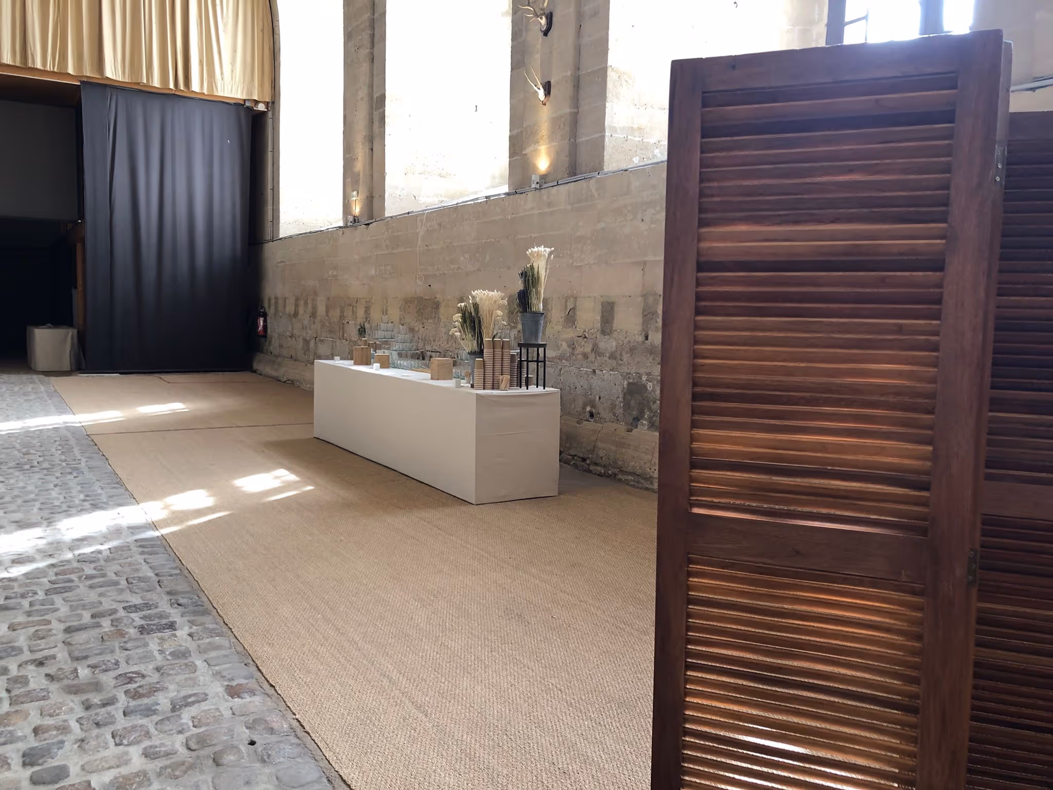 Indoor space with cobblestone and carpeted floor featuring a white table set with glasses, cups, and vases with dried plants, next to a large wooden folding screen.