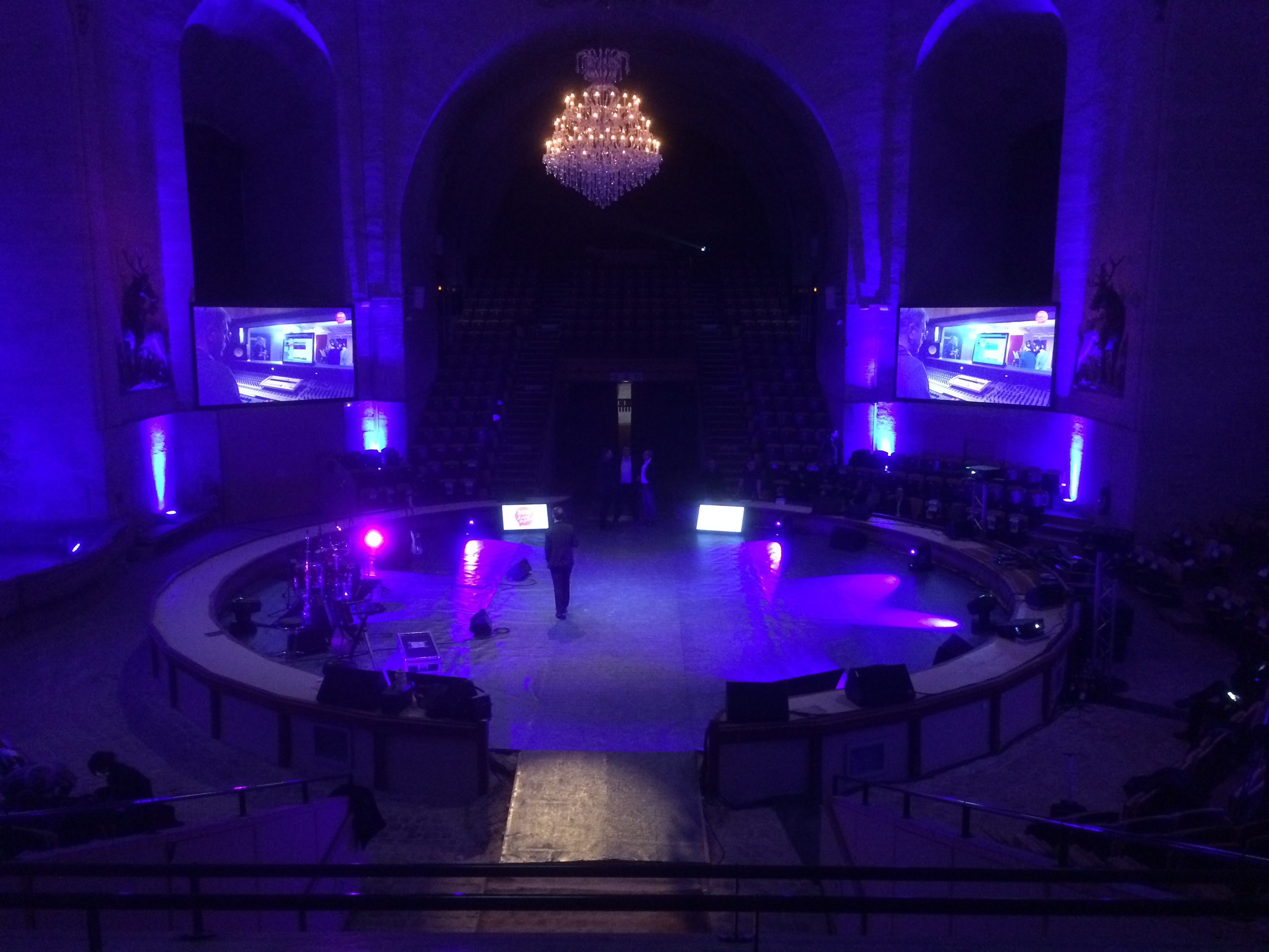 Dimly lit circular stage with purple lighting, a chandelier above, two large screens showing a man in a recording studio, and a few people standing on stage.