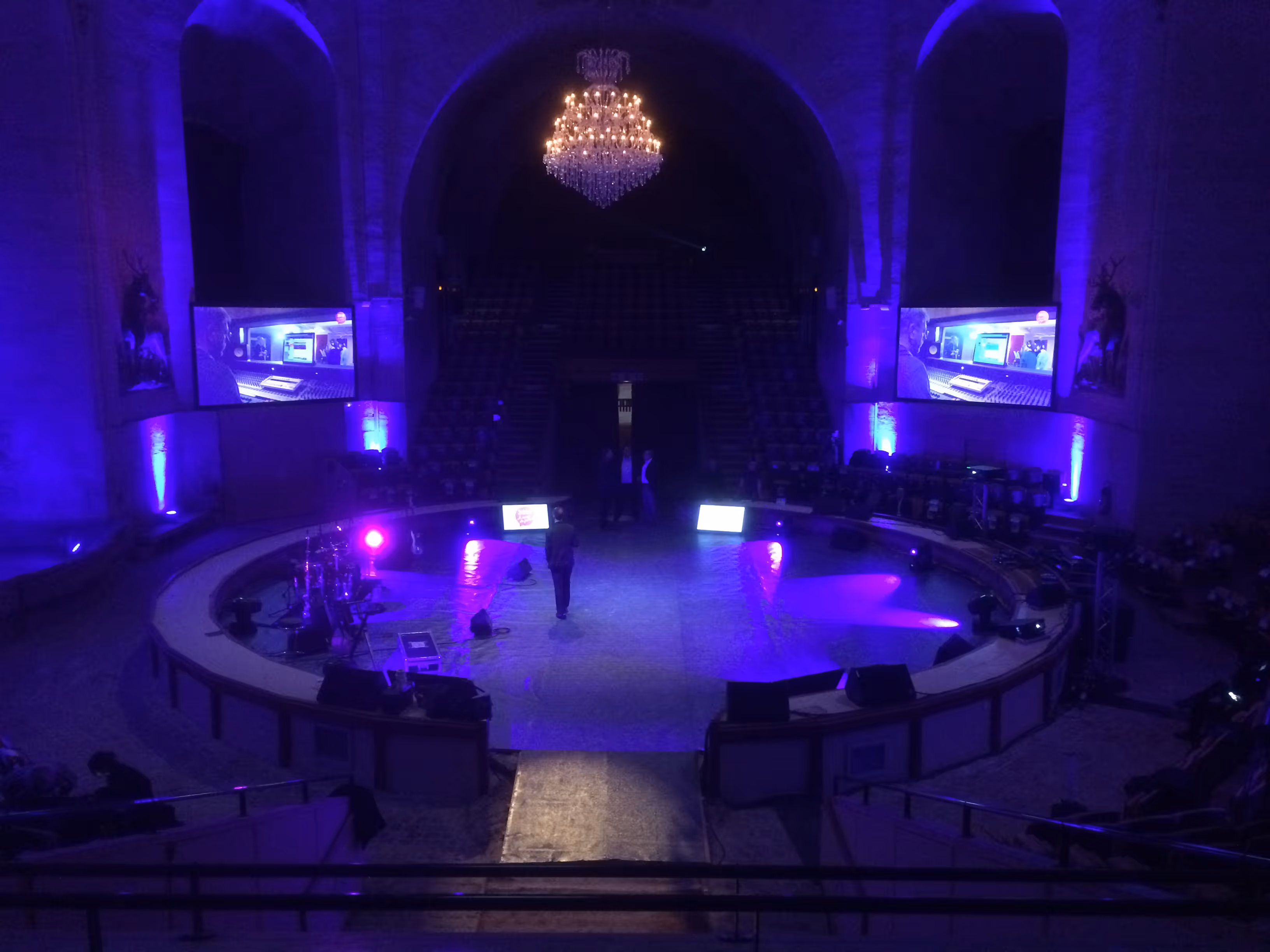 Dimly lit circular stage with purple lighting, a chandelier above, two large screens showing a man in a recording studio, and a few people standing on stage.