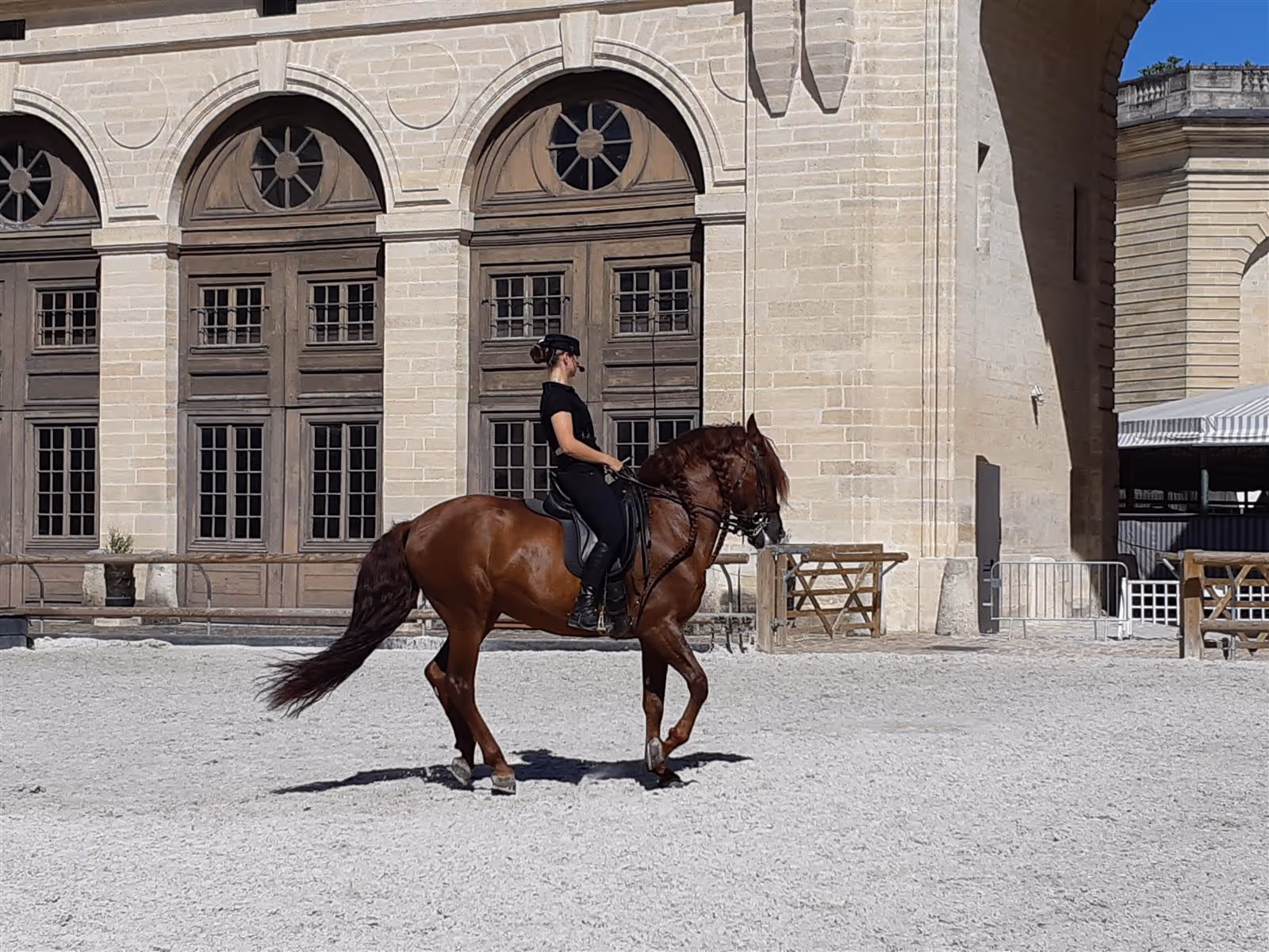 Equestrian rider in black attire performing dressage on a chestnut horse in front of a historic stone building.