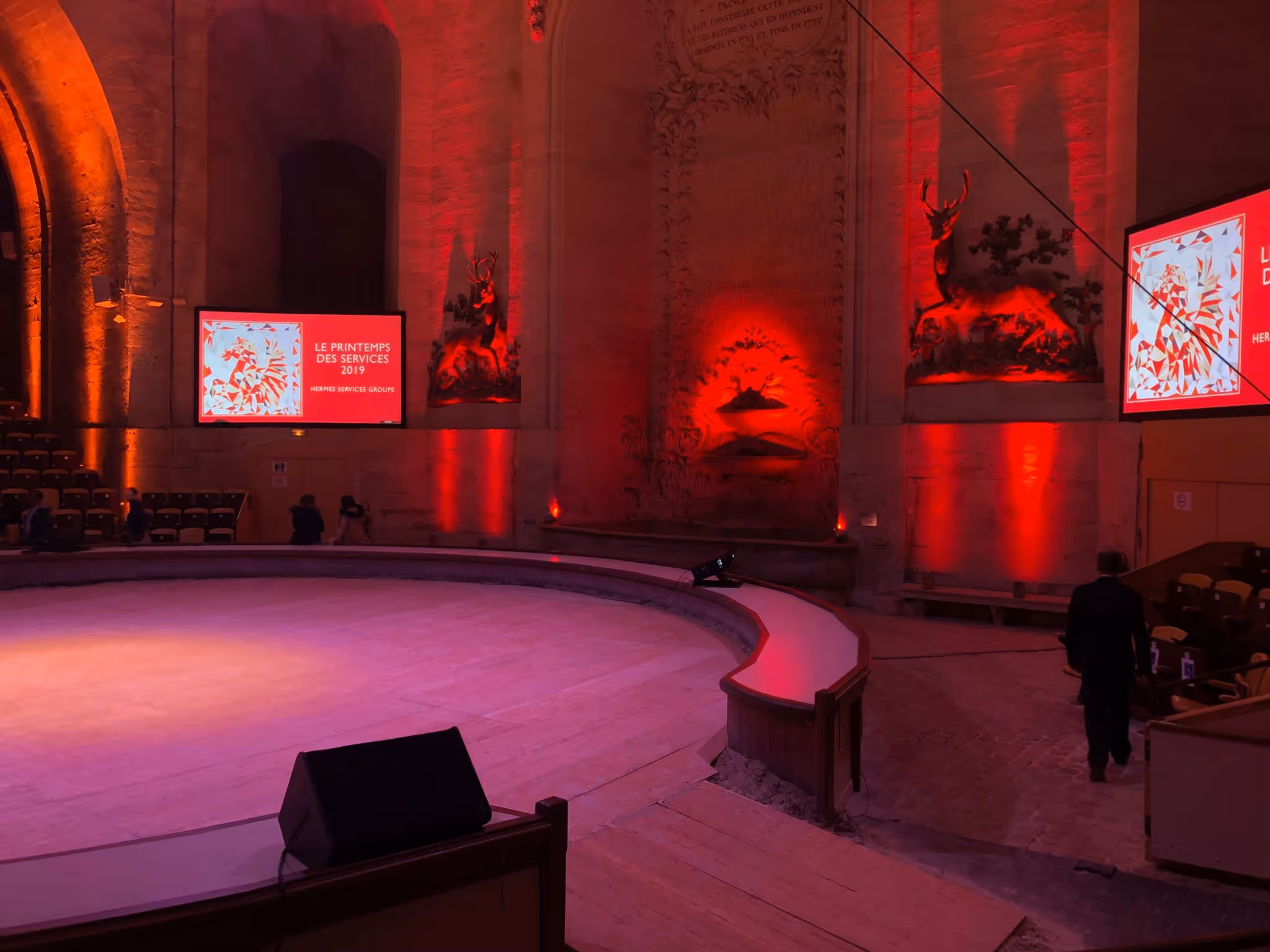 Interior of a large historic venue with red lighting, a round wooden stage, mounted deer sculptures, and screens displaying 'Le Printemps des Services 2019'.