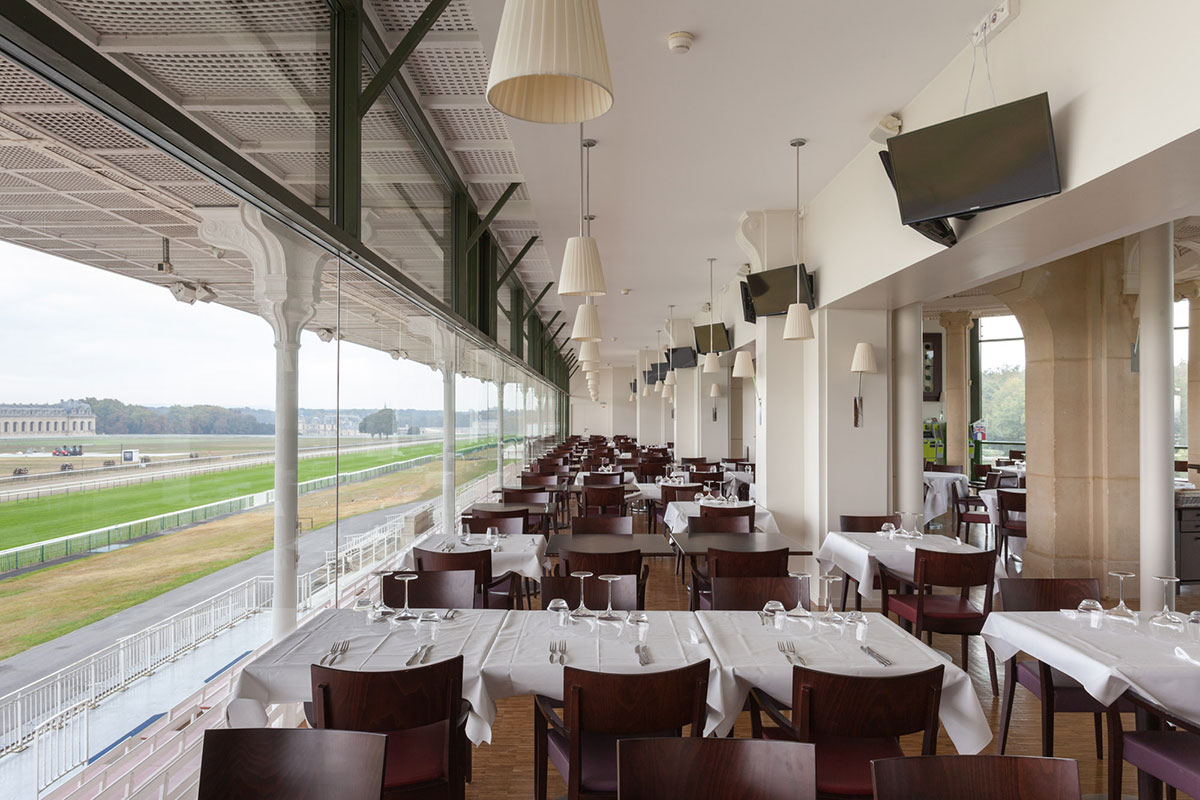 Indoor dining area with multiple tables covered in white tablecloths and wooden chairs, overlooking a racetrack through large glass windows.
