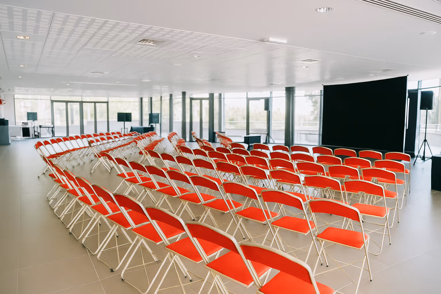 Empty conference room with rows of red folding chairs arranged facing a large black screen.