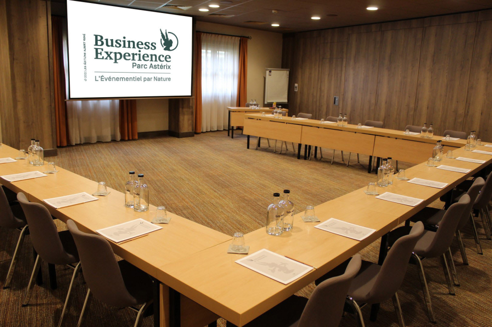 Conference room set up in a U-shape with wooden tables, grey chairs, water bottles, glasses, and papers, a projection screen displaying Business Experience Parc Astérix logo.