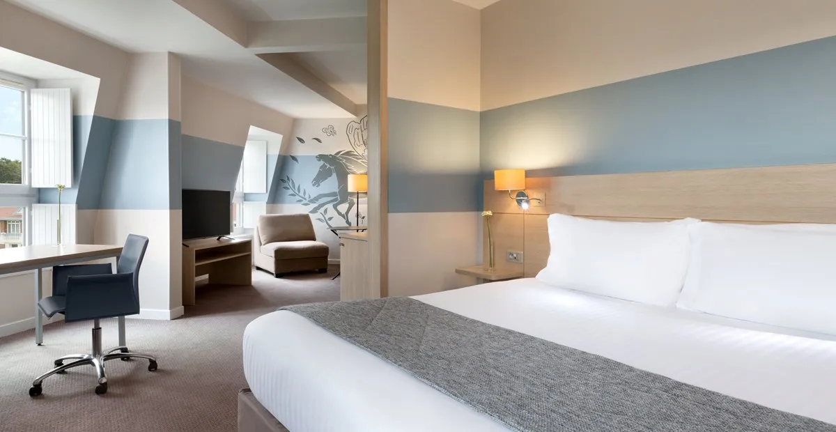 Bright hotel room with a large bed, gray and white bedding, blue and beige walls, a desk with a chair by the window, and a seating area with armchair and TV.