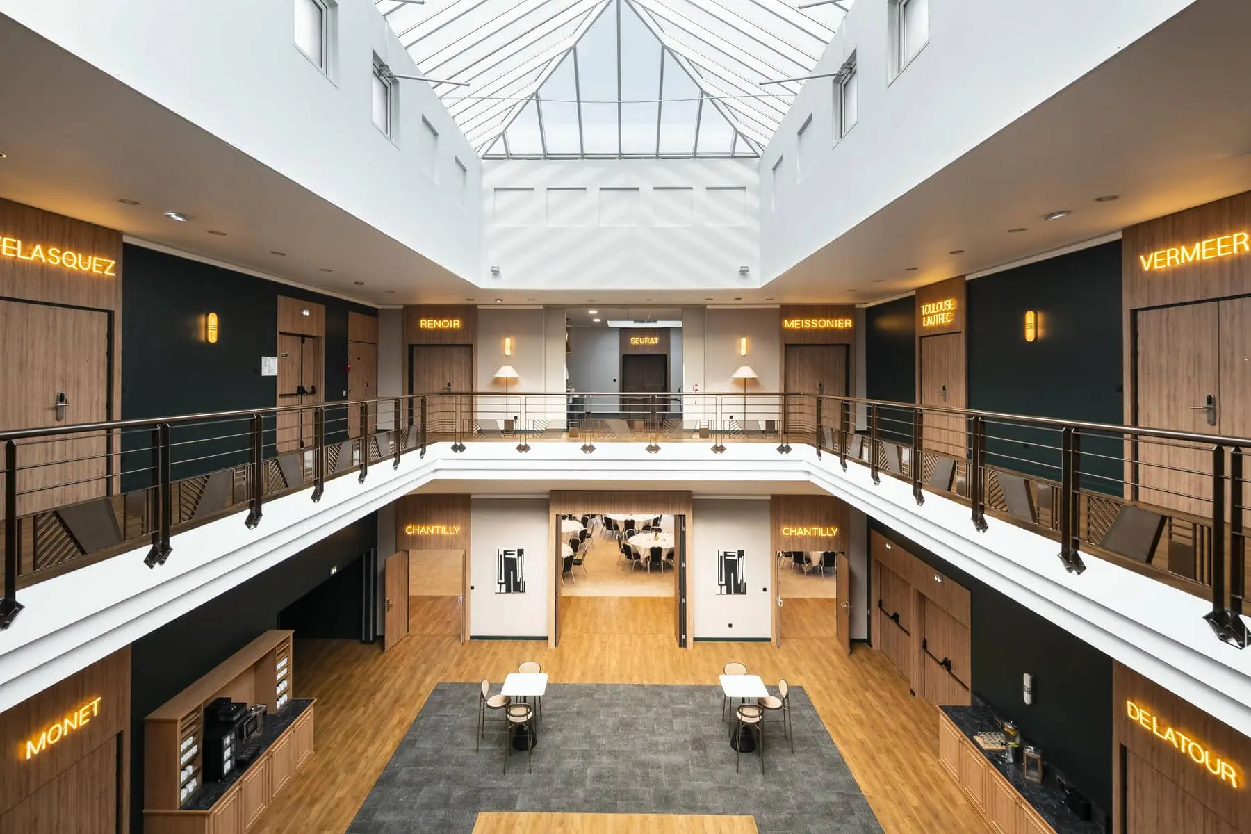 Modern two-story interior hallway with wooden doors labeled with famous painter names, a central skylight, and seating area with tables and chairs.