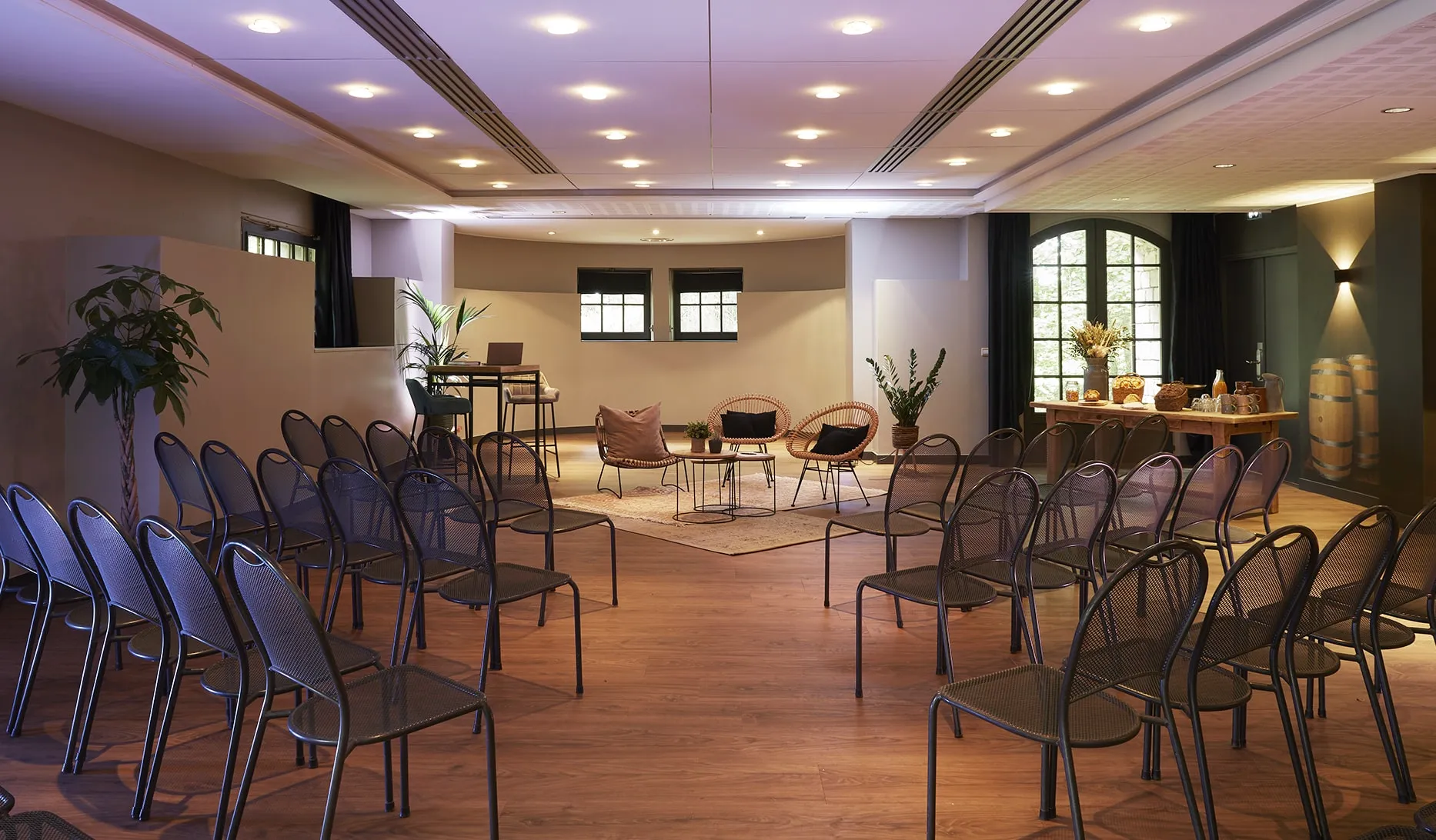 Modern seminar room with rows of black mesh chairs facing a seating area with wicker chairs and tables on a wooden floor.