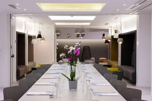 Modern conference room with a long white table set with water bottles, glasses, and notepads, surrounded by gray chairs, with purple orchids centerpiece and ambient ceiling lights.