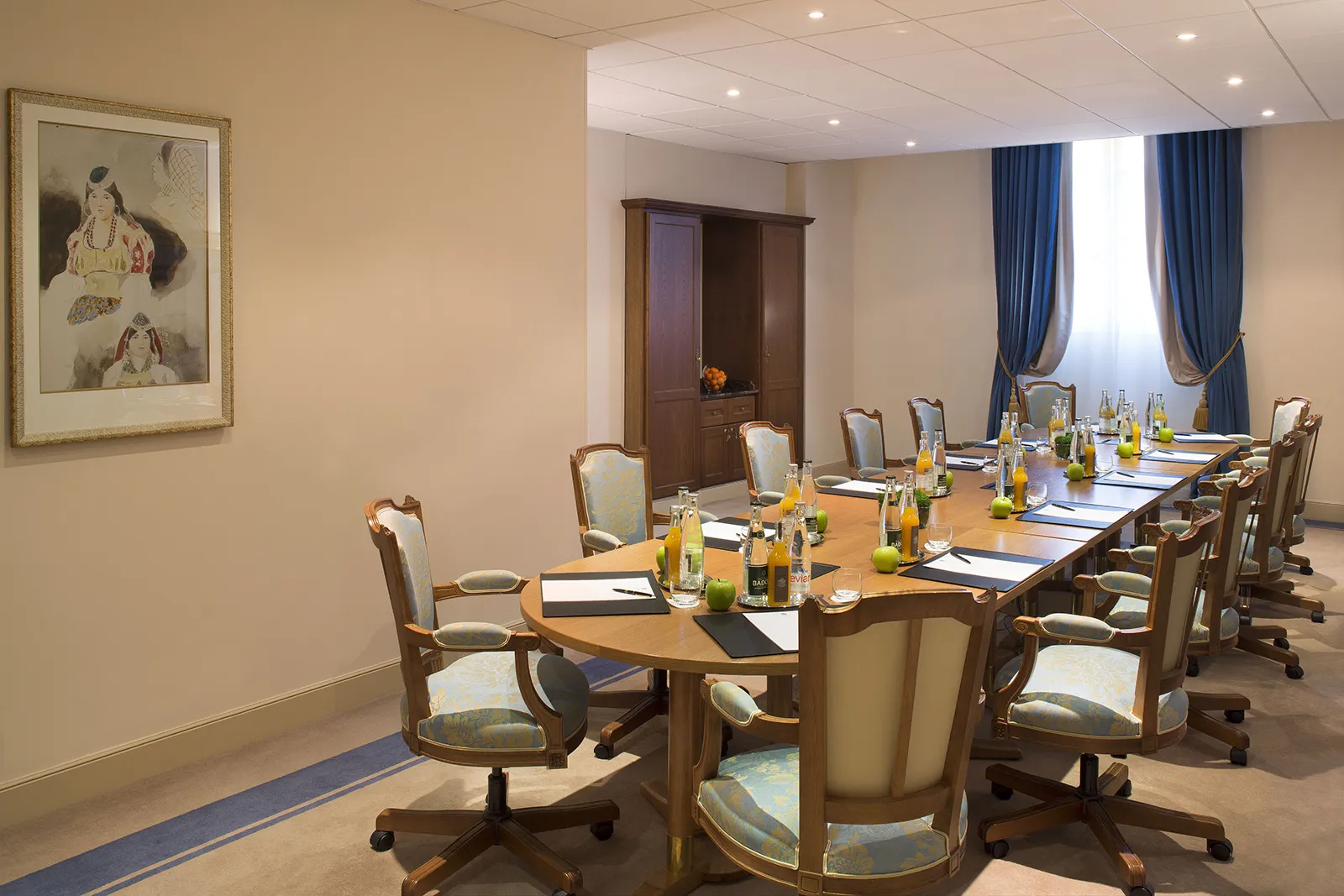Elegant conference room with a large wooden table, upholstered swivel chairs, notebooks, pens, bottled drinks, and green apples arranged for a meeting.