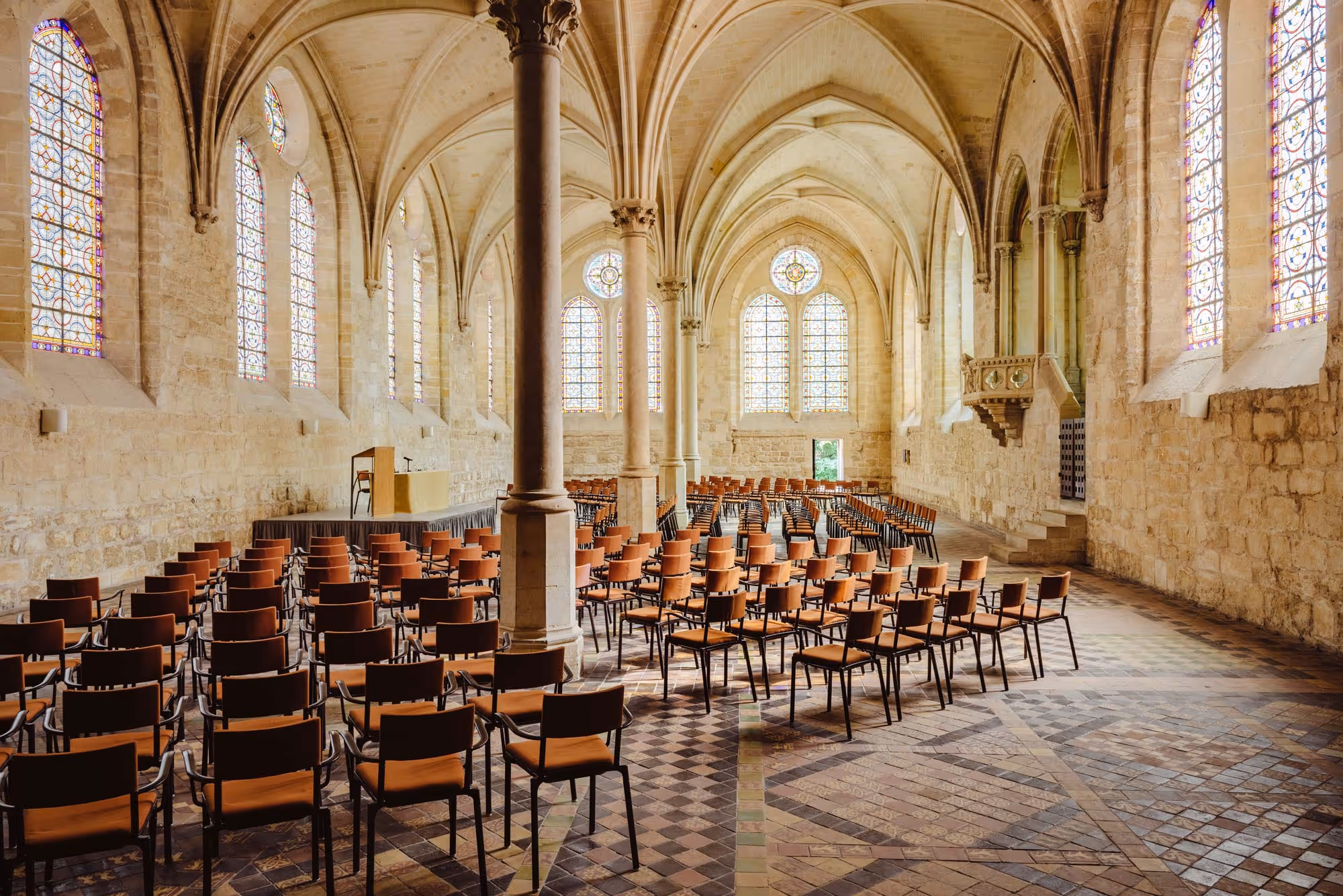 Interior of a large, historic stone chapel with stained glass windows and rows of brown chairs facing a podium.