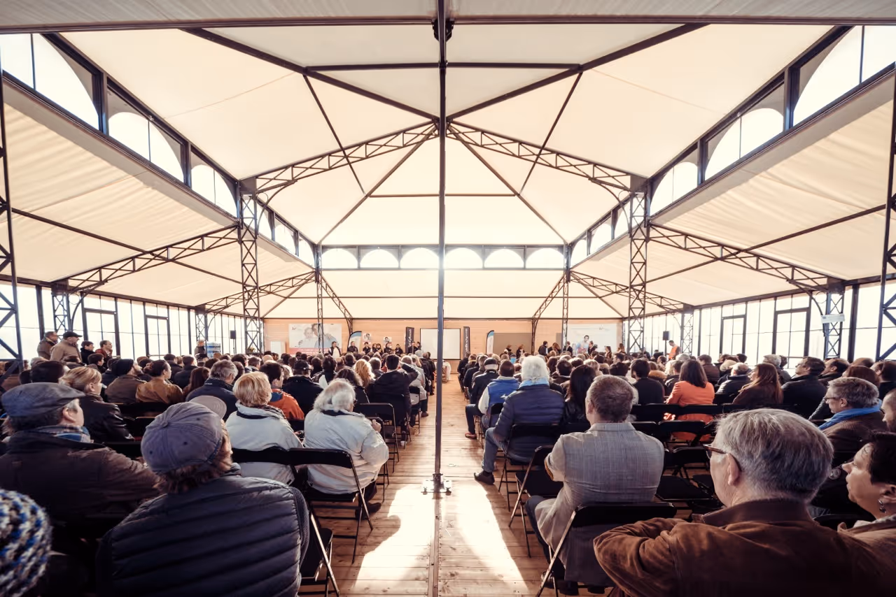 Large audience seated in rows inside a bright, metal-framed conference tent facing a stage.