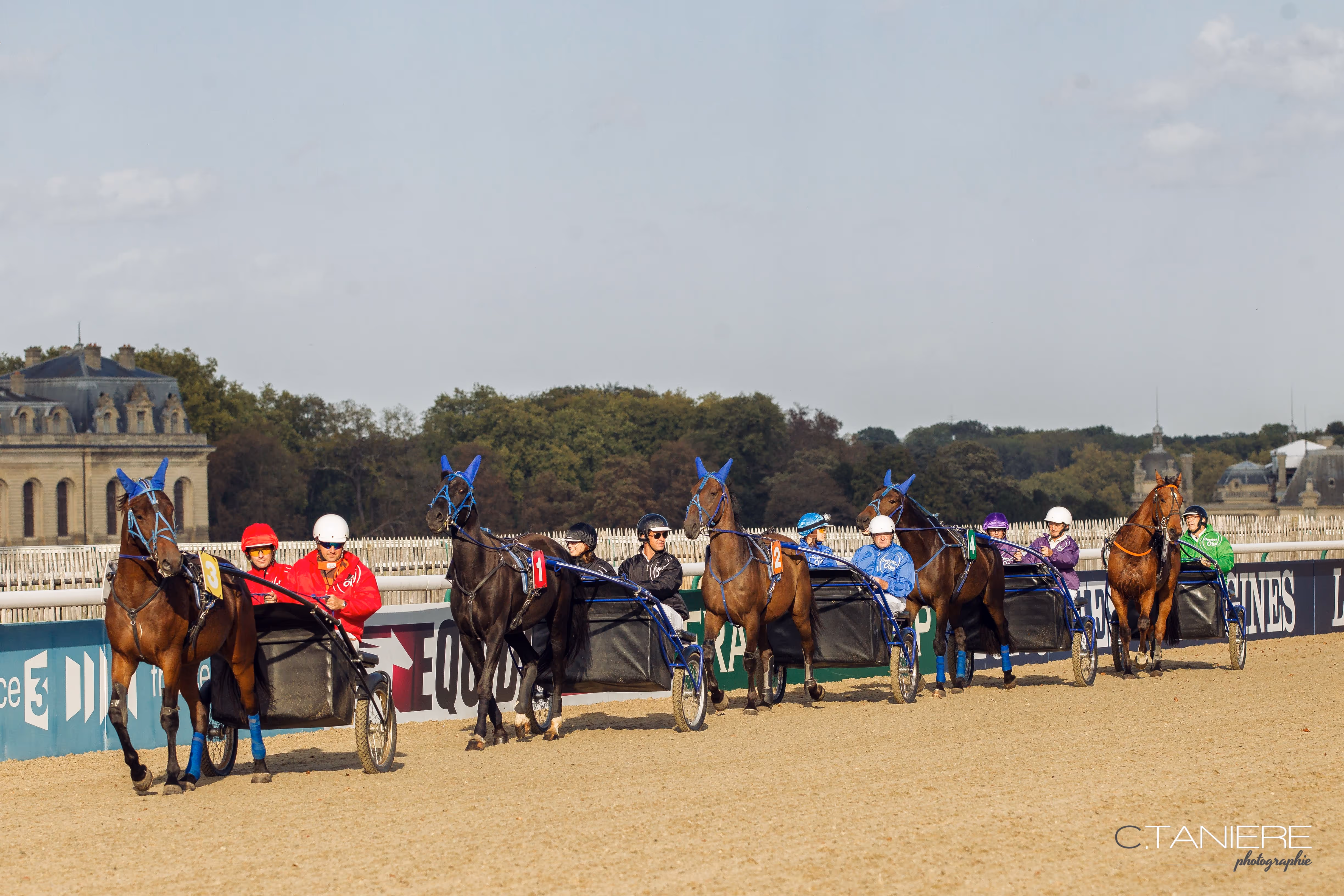 Five harness racehorses with drivers in colorful racing gear lined up on a dirt racetrack.